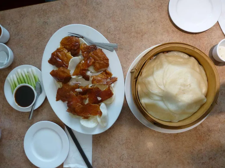 Platter of sliced Cantonese roast meats with shrimp chips, spring onions and dipping sauce, beside a bamboo steamer of steamed buns